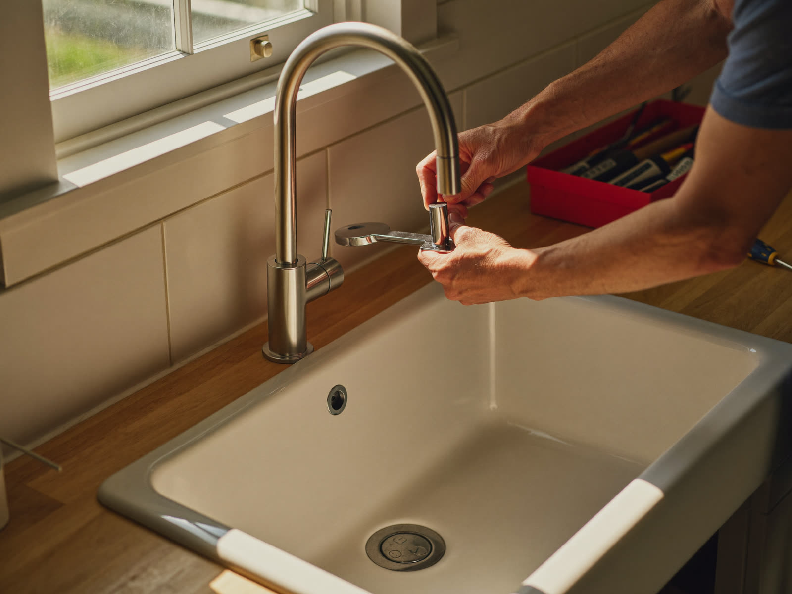 Skilled handyman hands installing a brushed-nickel kitchen faucet on a white farmhouse sink with a red toolbox in the background