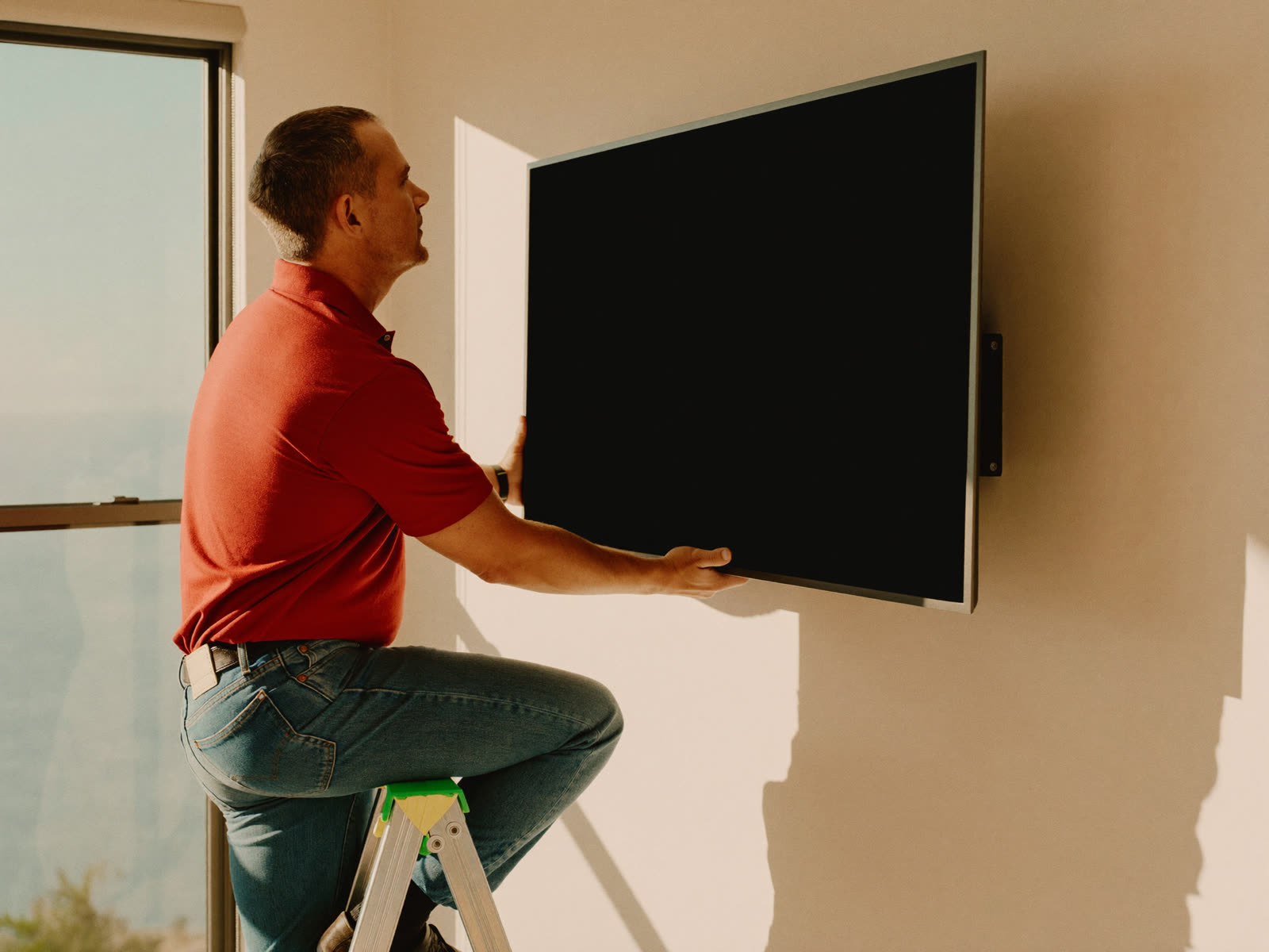 Hands holding a bubble level across a freshly-mounted black steel TV wall bracket, drill and drywall dust on the floor